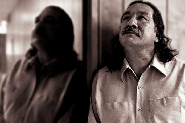 President Biden Commutes Sentence for Leonard Peltier Photo of a Native American man, Leonard Peltier, standing with his side against a mirror, gazing upwards.