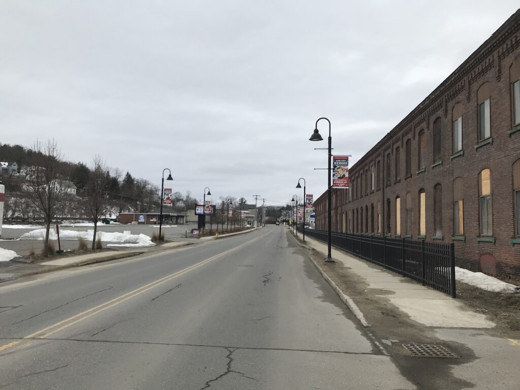Hometown Heroes banners hang from lampposts along both sides of a Fitchburg street beside a long brick building with boarded windows. Snow remains along the sidewalks.