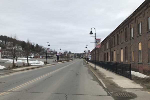 Hometown Heroes banners hang from lampposts along both sides of a Fitchburg street beside a long brick building with boarded windows. Snow remains along the sidewalks.