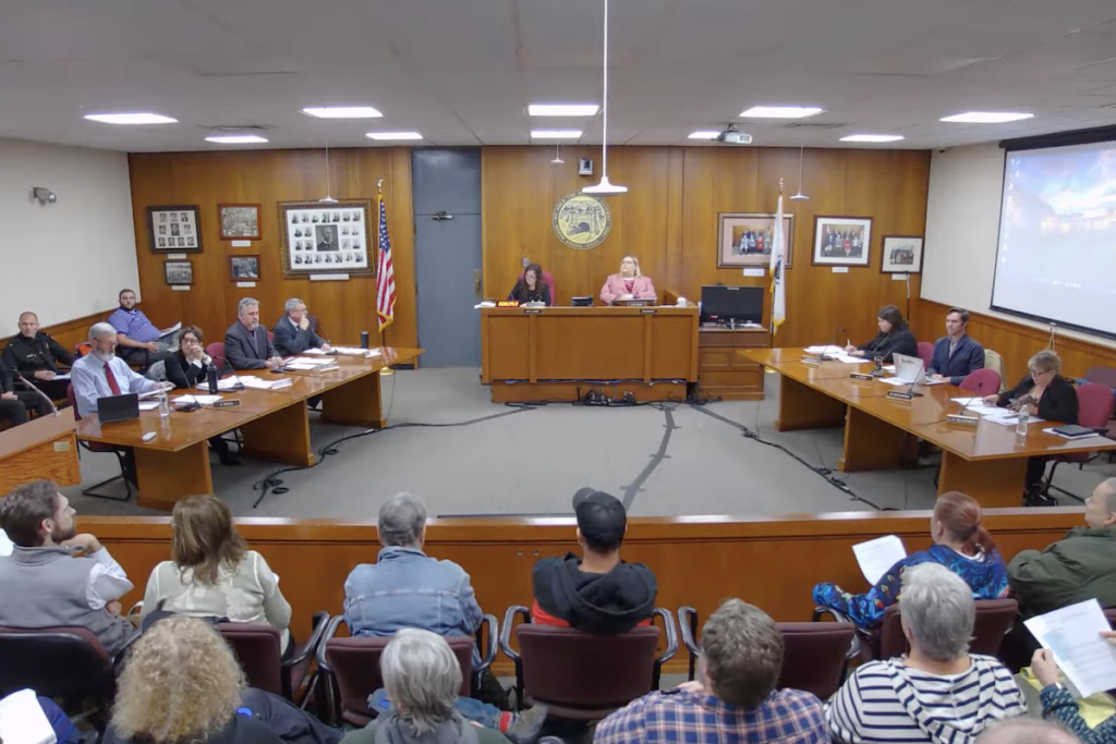 A full audience, seen from behind, faces North Adams city councilors seated in two rows on either side of Council President Ashley Shade in the wood-paneled council chambers.