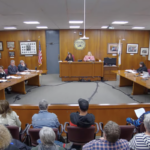 A full audience, seen from behind, faces North Adams city councilors seated in two rows on either side of Council President Ashley Shade in the wood-paneled council chambers.