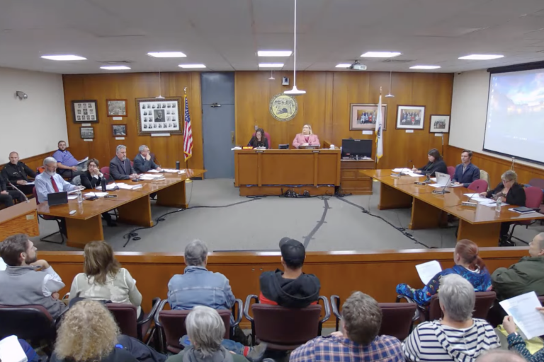 A full audience, seen from behind, faces North Adams city councilors seated in two rows on either side of Council President Ashley Shade in the wood-paneled council chambers.