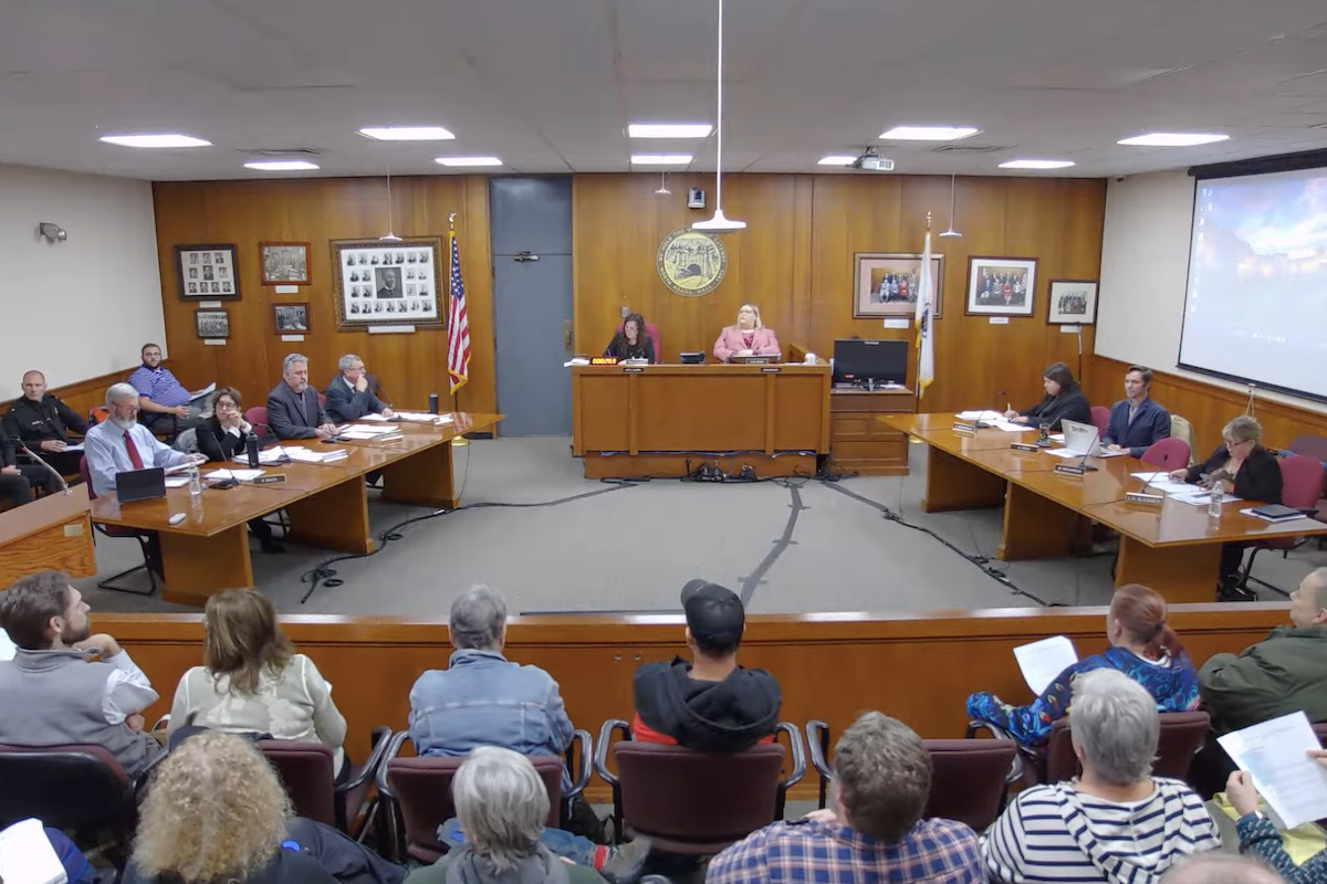A full audience, seen from behind, faces North Adams city councilors seated in two rows on either side of Council President Ashley Shade in the wood-paneled council chambers.